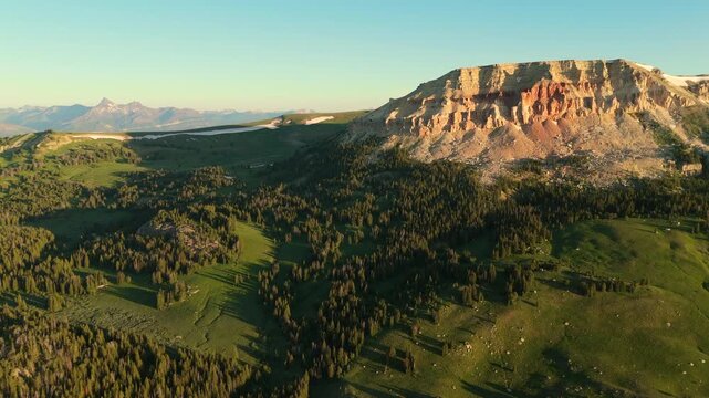 Beartooth Highway in Montana reveals stunning mountain landscape with red rock cliffs and lush green valleys. Pine forests stretch across rolling hills under golden evening light.
