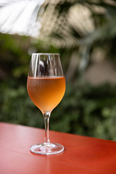 Glass of orange wine on bright red table with lush plants in the background in cafe or patio.