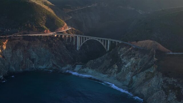 Aerial bixby bridge big sur coastline epic drone sweep over arch bridge hugging rugged cliffs and crashing waves golden hour light casting long shadows across coastal highway cinematic mood