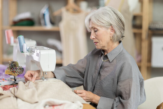 Elderly female seamstress sewing clothes on sewing machine in workshop
