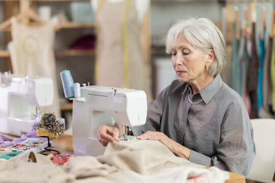 Skillful old tailoress sewing textile sitting at her workplace in tailor shop