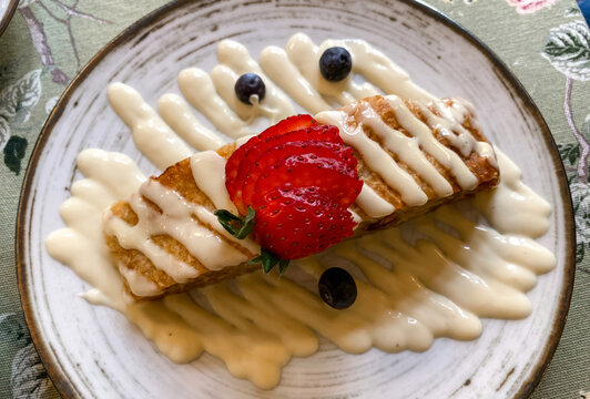 A delicious slice of fresh apple strudel served on a white plate with creamy vanilla sauce, topped with sliced strawberries and a blueberry