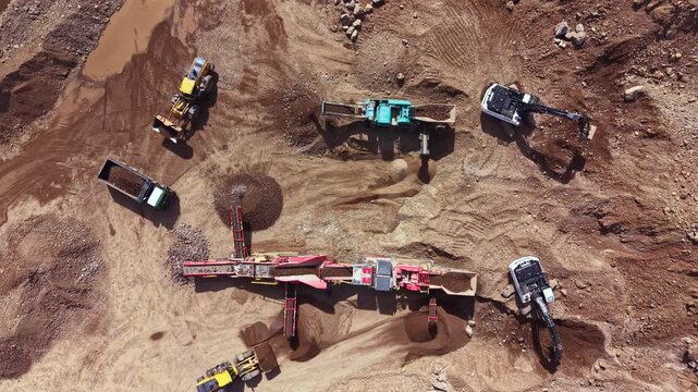 Large machines are operating on a mining site. Trucks and excavators are digging and transporting materials. The ground is dirt and rocks, with equipment arranged in a pattern.