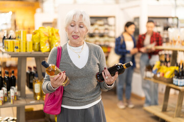 Naklejka premium Elderly woman buyer chooses bottle of wine in grocery store