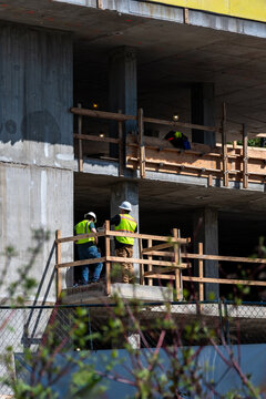 Construction workers in safety vests and hard hats standing on concrete floor of new build residential real estate development, condominium or apartment building, no walls, safety railings
