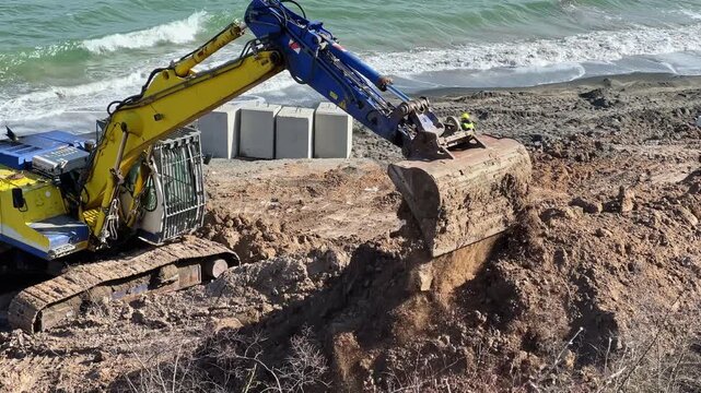 A large machine digs into the earth at a construction site near the beach. Waves crash in the background as workers prepare the area for development during a sunny day.