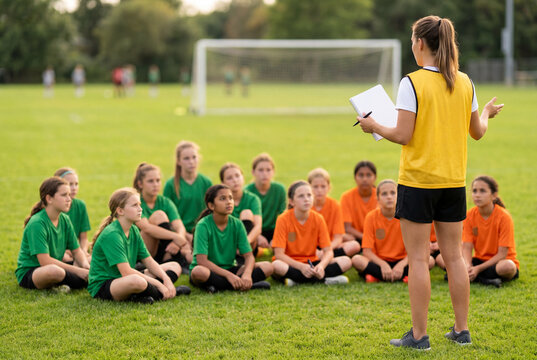Female soccer coach with clipboard instructing young girls sitting on green grass field