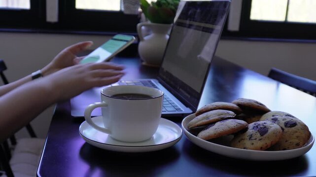 Female hands using smartphone while typing on laptop with coffee and cookies on desk.