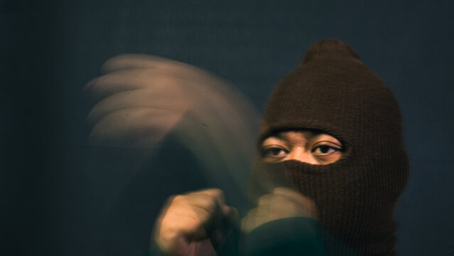 A male wearing a dark brown balaclava holds his fists up in a fighting stance with heavy motion blur trailing from his rapid arm movement. The dynamic and aggressive action builds