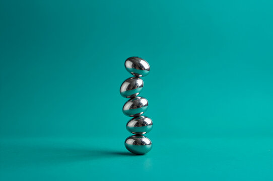 Stacked metallic ellipsoids form an unstable vertical column against a saturated teal backdrop, featuring high-contrast specular highlights and distorted chrome reflections.