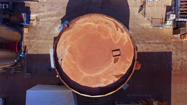 A large circular area holds a significant amount of sand in a construction site. Machinery and buildings surround this area. The sun is shining above during the daytime.