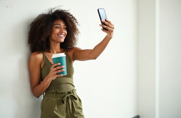 Fototapeta premium Smiling woman takes selfie with coffee cup in casual outfit. She holds phone in one hand, beverage in other, leaning on wall. Modern lifestyle and connection portrayed in portrait.