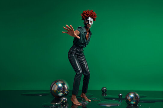 Avante-garde model with metallic silver facial mask and textured coily auburn hair poses amidst scattered chrome spheres on a dark reflective floor against an emerald green backdrop.