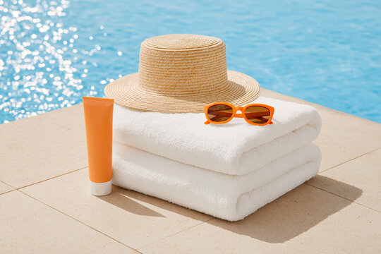 Summer poolside scene with straw hat orange sunglasses sunscreen tube and folded white towels placed on a stone surface near blue water