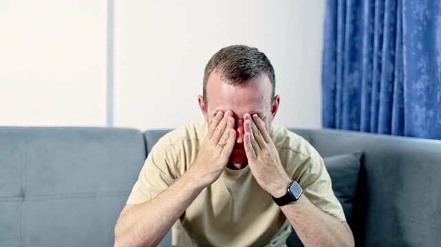 A man is sitting on a couch with his hands covering his face. He appears to be feeling stressed and is alone in a room with blue curtains and light coming in.