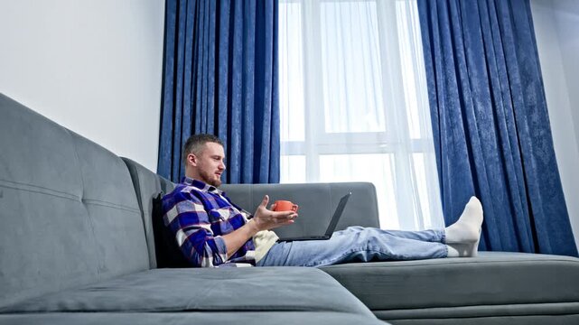 A man wears a plaid shirt while sitting on a gray sofa. He uses a laptop and looks focused. Sunlight enters through a window with blue curtains.