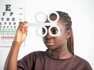 A young Black woman uses an optical flipper for a vision exam. A Snellen eye chart is blurred in the background. High-quality clinical photography focusing on eye health and optometry. © RODWORKS