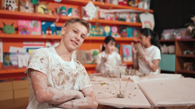 Smiling boy looking at camera and crossing arm with confident at workshop while diverse student having pottery class together. Happy caucasian student smile while pose with arm folded. Edification