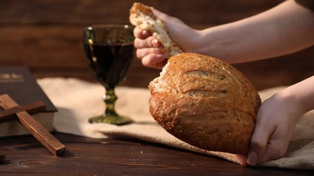 Sacrament of Communion. Woman breaking off loaf of bread at wooden table with glass of wine, cross and Bible, closeup