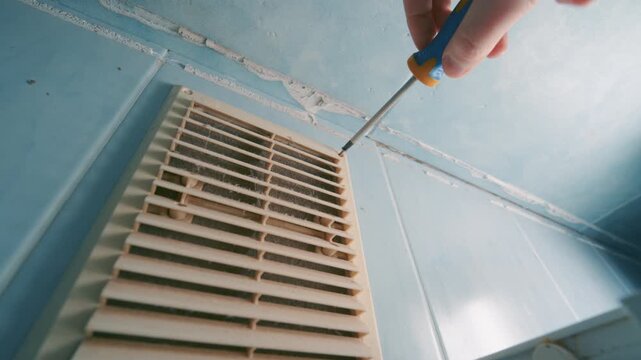Hand Using Screwdriver to Remove Dirty Ventilation Grille on Bathroom Wall | Home HVAC Maintenance and Air Vent Cleaning Close-Up | Indoor Repair Work