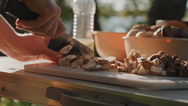 Camping food prep outdoors chopping wild mushrooms on cuttingboard, closeup hands slicing into small cubes beside bowl and bottle, sunlit trees and calm water beyond, warm golden hour light, rustic