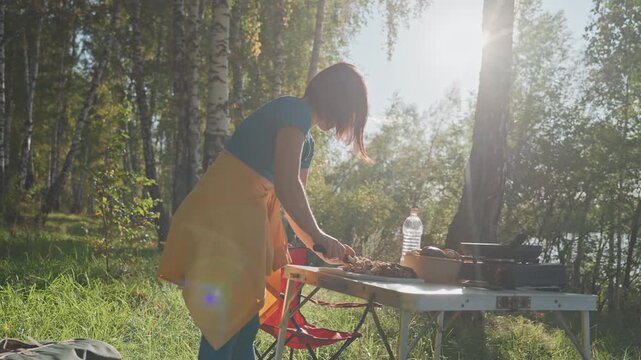 White woman arranging lakeside camping table birch trees golden sunlight. Portable stove and bottle rest on folding table, red chair tucked nearby, gentle breeze through leaves. Woman adjusts cookware