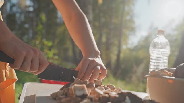 White woman slicing cooked meat campsite cutting board sunlight. Closeup hands guide knife through diced meat, bowl and water bottle visible, red chair background, focused culinary prep for outdoor