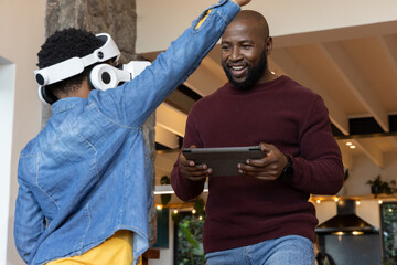 Naklejka premium African American males in kitchen, adult in sweater guiding denim-jacket teen using VR and tablet