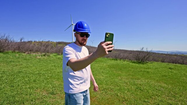 A man wearing a hard hat and sunglasses stands in a green field with wind turbines in the background. He holds a phone to take a selfie during a sunny day.