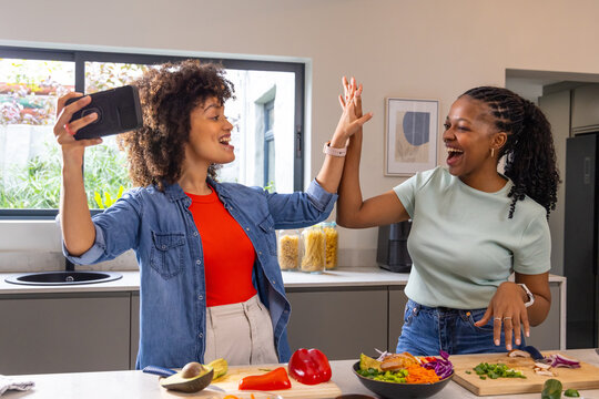 African American women friends high-fiving while preparing salad on kitchen island with smartphone