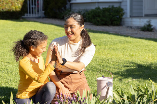 Asian mother and African American child laughing and high-fiving kneeling on lawn near watering can