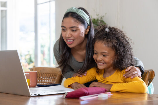 Asian mother and African American girl leaning and studying at wooden table with laptop, pencil