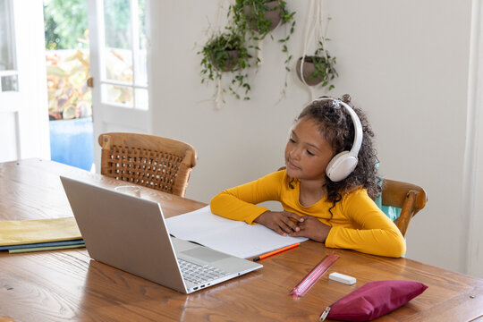 African American girl child working at dining table wearing yellow top with laptop, headset, pencil