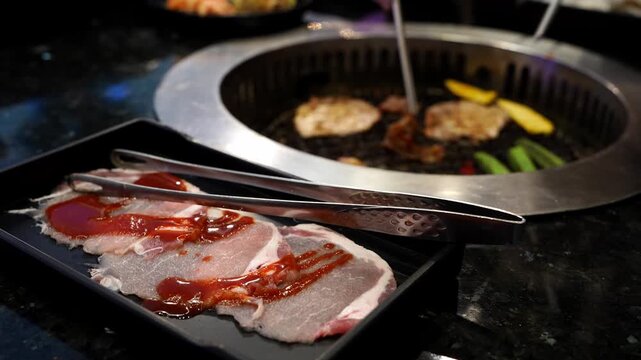 Raw sliced meat with savory sauce on a black tray and metal tongs in a Korean barbecue restaurant. Grilling meat and vegetables are visible in the background on the built-in stovetop grill.