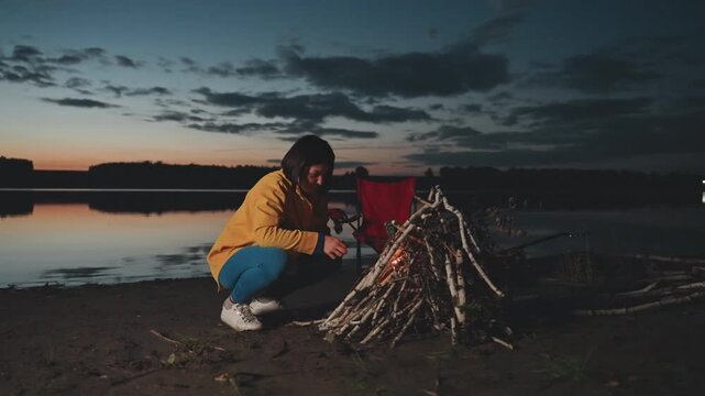 Twilight camping ignite by lake, solo camper kneeling adding tinder and match, sparks catch, red chair behind, quiet shoreline mood, rising flame brings warmth and excitement to microadventure