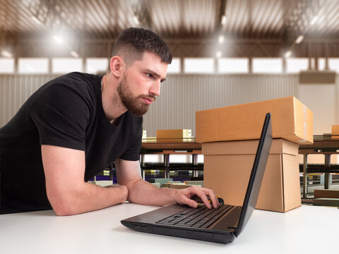 Warehouse operator uses laptop beside parcels, processing online orders and shipment logistics.