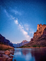 Fototapeta premium Stunning view of starry night sky with vibrant Milky Way over illuminated rocky canyon cliffs reflecting in calm river water at dusk