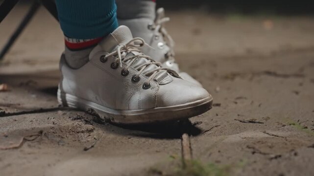 Closeup silver sneaker on campsite ground, muddy texture around sole, colorful socks visible, camper sitting on folding chair, casual relaxed pause before hike, soft morning light and scattered grass