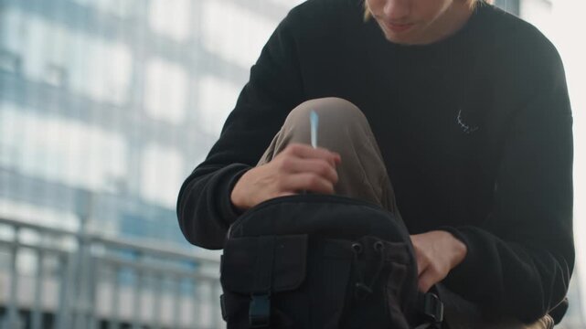 Youth checks gadgets. Young individual inspecting tiny equipment attentively in morning light. Youth crouched beside barrier reviewing compact gadget under soft dawn rays in city background