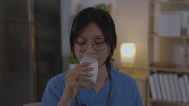 Tired asian doctor woman in relaxed moment at night, drinking glass of milk for good sleep. This healthcare professionals quiet wellness routine after long day