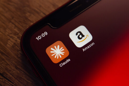 Mexico City, MEXICO - Apr 27 2026 : Angled close-up of an iPhone showing the Claude and Amazon app icons against a dark red background on a wooden surface.
