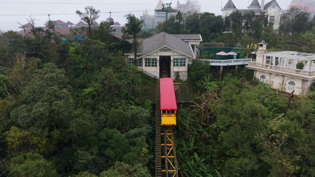 Vertical screen, Vietnam, Da Nang, Ba Na Hills Aerial View Red Cable Car Approaching Station With Lush Forest And Colonial Buildings, Slow Ascent On Yellow Steel Track, Maintenance Worker