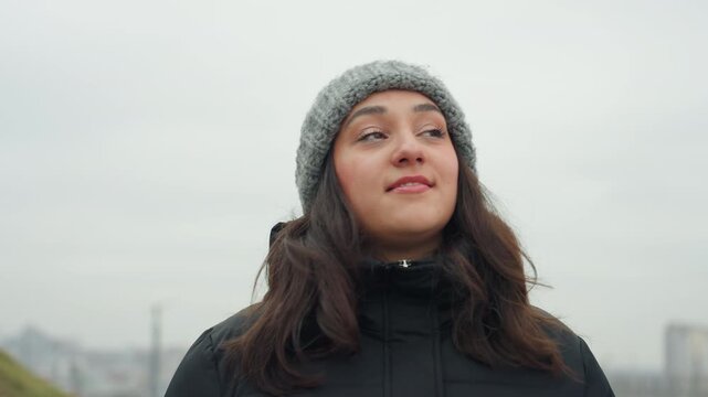 Bright young lady smiling while admiring sky in urban surroundings during chilly conditions. Content woman gazing happily skyward amidst bustling cityscape on cold day with cheerful expression