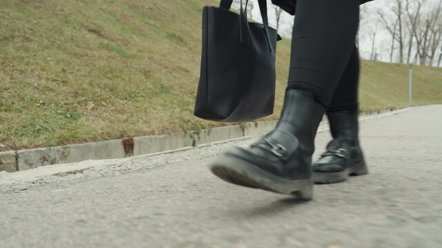 Low angle white woman closeup feet walking on pavement percussive footwork and bag swing create rhythmic pulse focus on boots, toe and heel contact with curb detail gritty concrete texture and urban