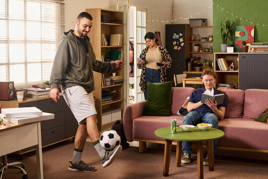 Young adult roommates studying and relaxing in shared apartment, man juggling soccer ball while woman checking smartphone and man reading book. Ideal for student housing, campus lifestyle marketing