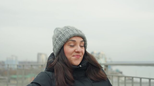 South asian woman dancing on rooftop, spinning arms and smiling, wearing gray beanie and black puffer coat, urban skyline background, cold overcast day, playful wind lifts hair, candid portrait