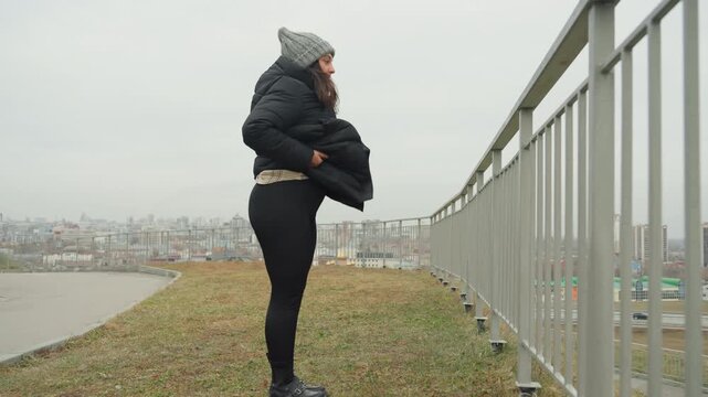City overlook dance warmup railing view, solo dancer practicing leg extension in puffer coat and knit beanie, high kick to railing then seated stretch, overcast skyline and muted winter light, playful