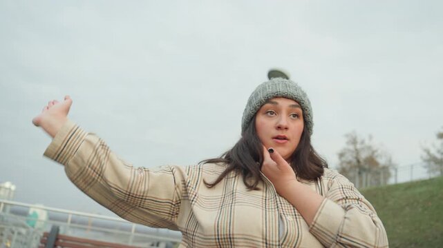 Closeup balcony dance woman in knit hat showing expressive hand gestures and slow choreography against overcast sky bench, lamp, and railing frame scene introspective mood, solo dancer practicing