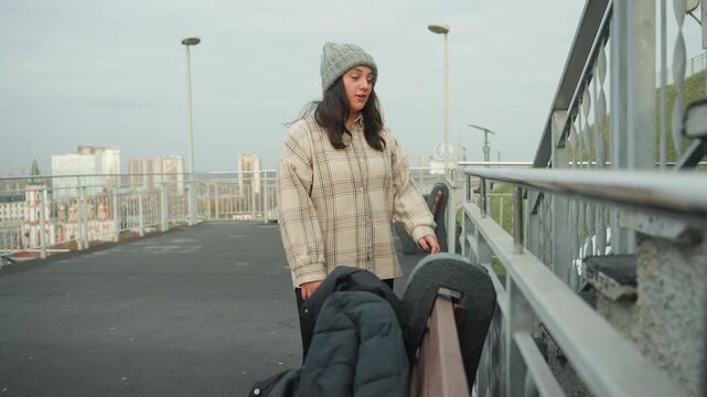 White woman playful dance on bridge with chunky sweater and knit beanie, skyline backdrop and overcast light. Frame commuter crossing with purposeful step. Frame impromptu performer breaking into