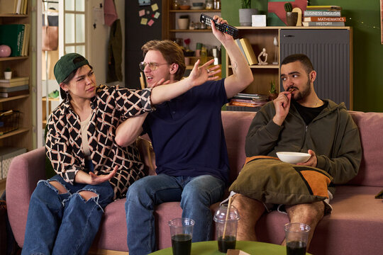 Young adult friends arguing over television remote while watching program on couch, expressing conflict in shared living. Useful for student lifestyle, roommate tension, entertainment habits